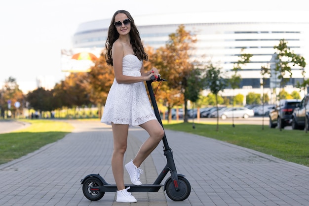 Dziewczyna w letniej sukience jeździ skuterem elektrycznym po chodniku