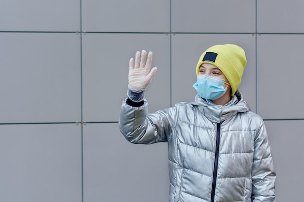 Zdjęcie dziewczyna w masce medycznej i rękawiczkach ochronnych na zewnątrz, machając ręką, witając kogoś.