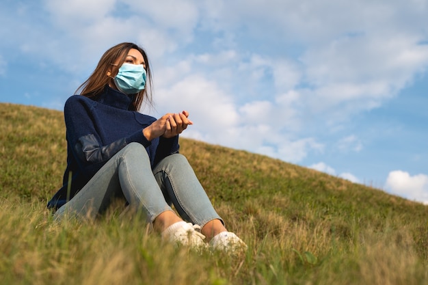 Dziewczyna w masce medycznej siedzi na zielonej trawie na tle błękitnego nieba