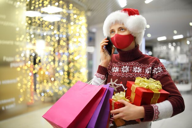 Dziewczyna w masce trzyma prezenty i torby rozmawia przez telefon w centrum handlowym przed Świętami Bożego Narodzenia