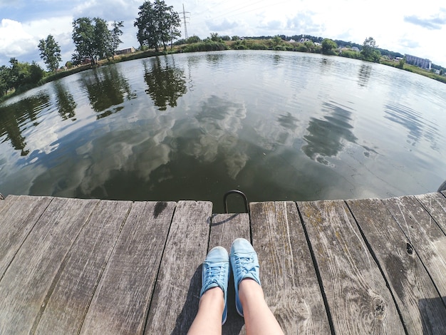 Dziewczyna w niebieskich butach siedzi na drewnianym molo w pobliżu rzeki jeziora