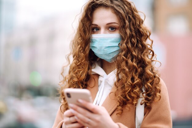 Dziewczyna w ochronnej sterylnej masce medycznej na twarzy z telefonem w mieście kwarantanny. Kobieta szuka wiadomości za pomocą telefonu.