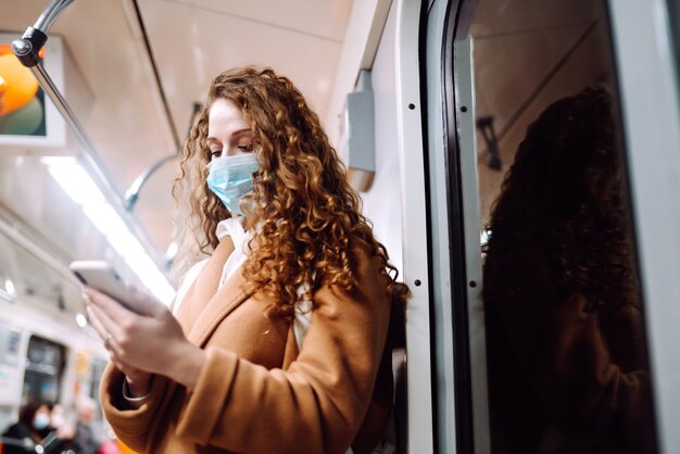Dziewczyna w ochronnej sterylnej masce medycznej na twarzy z telefonem w wagonie metra. Kobieta wyszukująca informacje o koronawirusie przez telefon. Koncepcja zapobiegania rozprzestrzenianiu się epidemii.