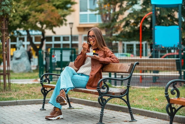 Dziewczyna w okularach i modnych ubraniach siedzi na ławce w parku i zajada pyszne słodycze Selektywne skupienie