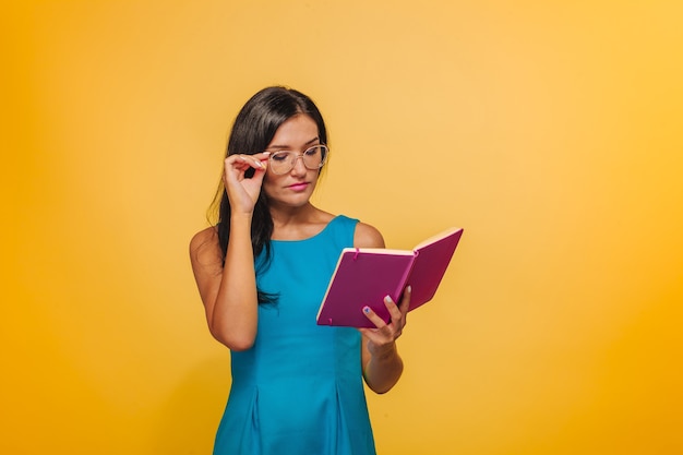 Dziewczyna w okularach na żółtym tle w niebieskiej sukience czyta różowy notatnik