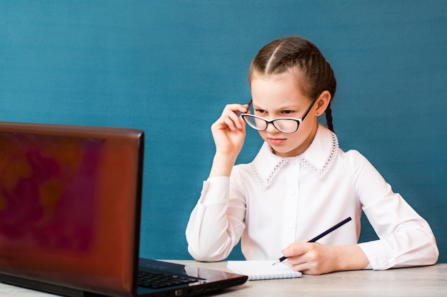 Dziewczyna w okularach pracuje przy laptopie i robi notatki w zeszycie przy stole