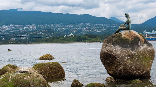 Dziewczyna w piance rzeźba z brązu siedzi na skale w wodzie morskiej i panoramę miasta Vancouver w Kanadzie