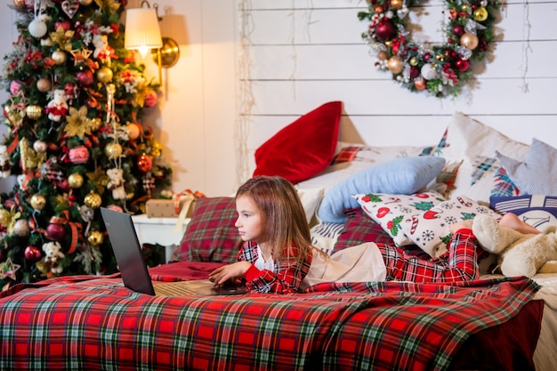 Dziewczyna w piżamie leży na łóżku na czerwonej kratce choinki i patrzy na monitor laptopa.