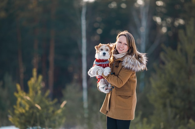 Dziewczyna w płaszczu trzyma w ramionach psa Jack Russell Terrier w zimowym lesie świerkowym na śnieżnej koncepcji Bożego Narodzenia