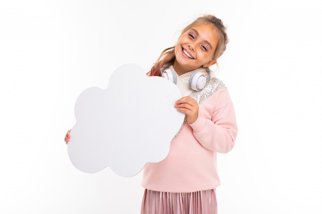 Dziewczyna w różowym swetrze z białymi słuchawkami na szyi na białej izolowanej powierzchni, dziecko trzyma pustą chmurę w dłoniach do tekstu