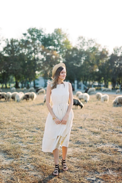 Dziewczyna w słomkowym kapeluszu stoi na trawniku z pasącymi się owcami