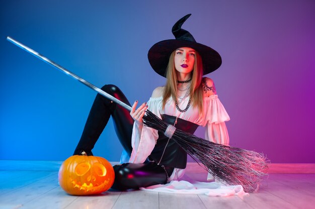 Dziewczyna w stroju wiedźmy na Halloween z miotłą i dynią w neonowym świetle