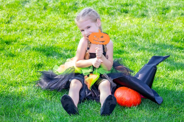 Dziewczyna w stroju wiedźmy na święta Halloween. Szczęśliwa dziewczyna siedzi na trawie z dyni0