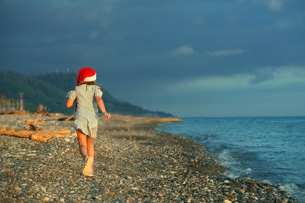 Dziewczyna w sukience w paski i czerwonej czapce Mikołaja biegnie do nas plecami brzegiem morza