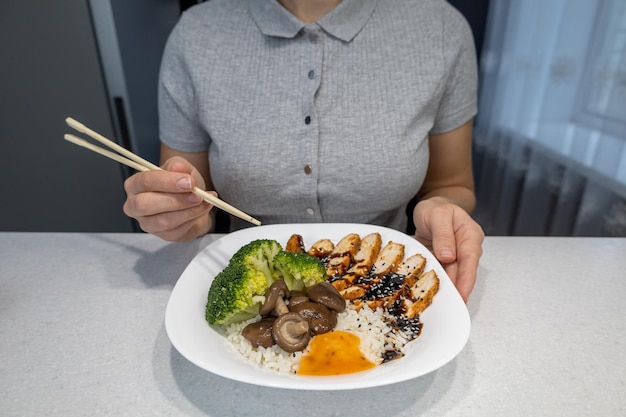 Dziewczyna w szarej koszuli w kuchni w domu trzyma w rękach talerz złotego ryżu, grzybów shiitake, kurczaka z sosem unagi i brokułami Koncepcja azjatyckiego jedzenia