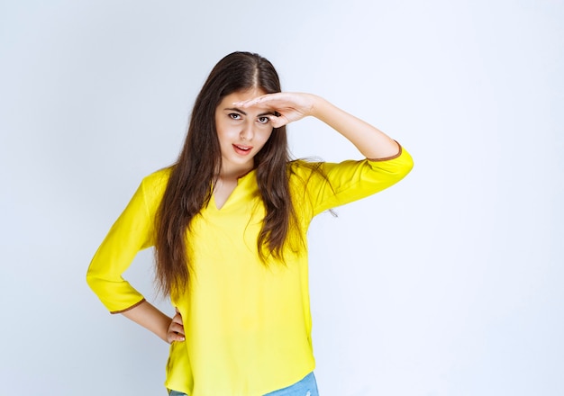 Dziewczyna w żółtej koszuli przykłada rękę do czoła i obserwuje lub szuka kogoś.