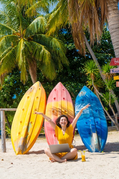 Dziewczyna w żółtej sukience na tropikalnej piaszczystej plaży pracuje na laptopie w pobliżu kajaków i pije świeże mango. Praca zdalna, udany niezależny. Działa na wakacjach.