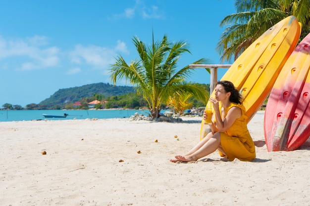 Dziewczyna w żółtej sukience na tropikalnej piaszczystej plaży pracuje na laptopie w pobliżu kajaków i pije świeże mango. Praca zdalna, udany niezależny. Działa na wakacjach.