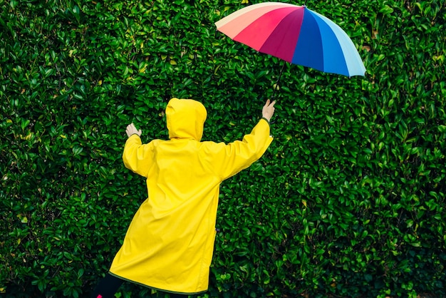 Dziewczyna w żółtym płaszczu przeciwdeszczowym na tle ściany z trawą z wielobarwnym parasolem