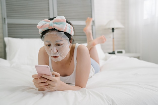 Dziewczyna z arkuszem maski urody relaks w domu sypialnia wiadomości tekst na telefon komórkowy. azjatycka Japonka przy użyciu smartfona w łóżku, czekając na zabieg na twarz. Koncepcja pielęgnacji kobiecej skóry i ciała.