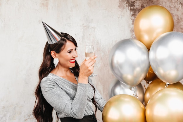 Dziewczyna z balonami śmieje się na tle ozdobnej ściany Piękna szczęśliwa kobieta na urodziny sylwestrowej imprezy świątecznej bawi się trzymając kieliszek szampana