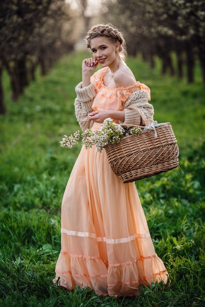 Dziewczyna z blond włosami w lekkiej sukience w kwitnącym ogrodzie. dziewczyna w pięknej sukience i swetrze z dzianiny cieszy się zachodem słońca w ogrodzie kwitnienia gruszy, z koszem kwiatów