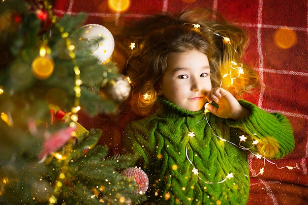 Dziewczyna z długimi włosami i girlandami leży na czerwonej kracie pod choinką z zabawkami w ciepłym swetrze z dzianiny. Boże Narodzenie, Nowy Rok, dziecięce emocje, radość, oczekiwanie cudu i prezenty