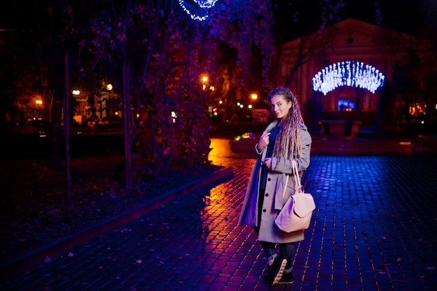 Dziewczyna z dredami spacerująca nocną ulicą miasta przed girlandowymi światłami