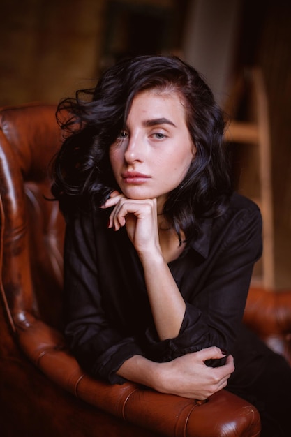 Dziewczyna z kręconymi ciemnymi włosami i naturalnym makijażem siedząca na skórzanym krześle w stylu vintage przetwarzania zdjęć