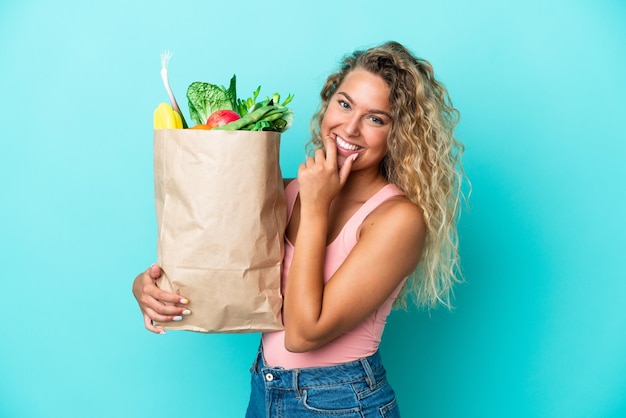 Dziewczyna z kręconymi włosami trzymająca torbę na zakupy spożywcze na zielonym tle szczęśliwa i uśmiechnięta