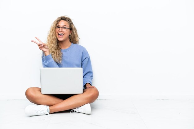 Dziewczyna z kręconymi włosami z laptopem siedząca na podłodze uśmiechnięta i pokazująca znak zwycięstwa