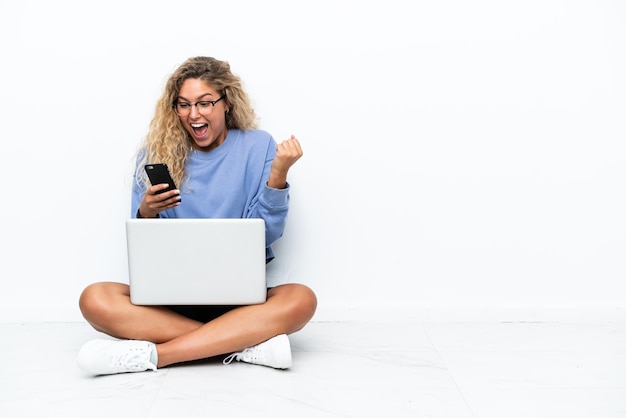 Dziewczyna z kręconymi włosami z laptopem siedząca na podłodze zaskoczona i wysyłająca wiadomość