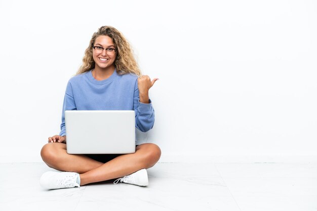 Dziewczyna z kręconymi włosami z laptopem siedzi na podłodze i wskazuje na bok, aby zaprezentować produkt