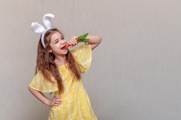 Dziewczyna z króliczymi uszami na głowie w żółtej sukience gryzie marchewkę.