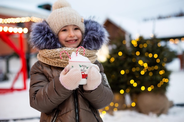 Dziewczyna z kubkiem z trzciny cukrowej i napisem Merry and Bright w jej rękach odkryty w ciepłych ubraniach w zimie na świątecznym targu Fairy lights girlands zdobione miasto śniegu na nowy rok Boże Narodzenie