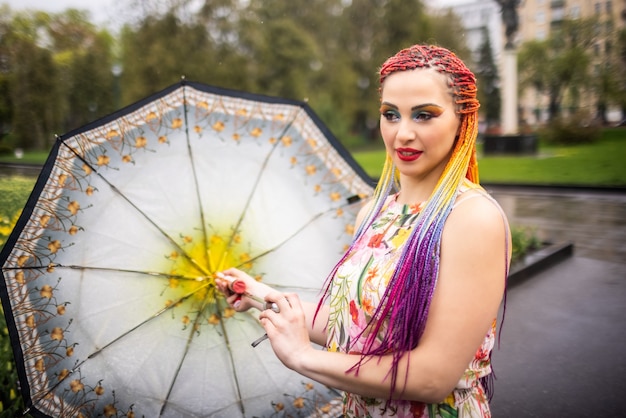 Dziewczyna z niestandardowym makijażem brokatowym z wielokolorowymi sztucznymi warkoczami w sukience w kwiatowy print. Zawiruj z parasolem w dłoniach na spacerze w wiosennym parku na tle kwiatów.
