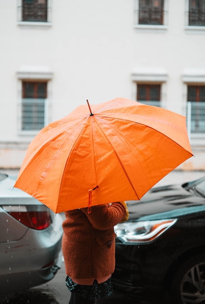 Dziewczyna z pomarańczowym parasolem chroniąca się przed deszczem spacerująca między samochodami