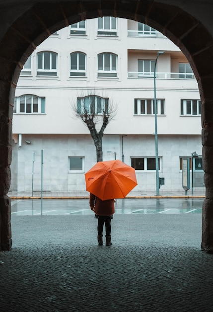 Dziewczyna z pomarańczowym parasolem stoi nieruchomo pod łukiem, obserwując, jak pada w mieście