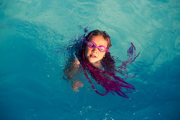 Dziewczyna z różowymi warkoczykami w różowych okularach pływackich pływa w basenie w ośrodku
