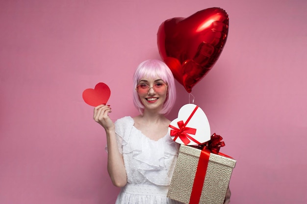 Dziewczyna z różowymi włosami w sukience trzyma prezenty i balony i pokazuje walentynkowe serce