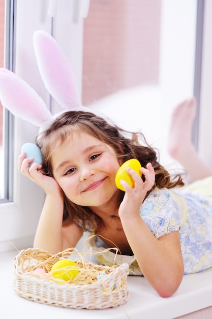Dziewczyna z uszami królika z pisanki w ręce uśmiecha się. wielkanocny koszyk siedzi przy oknie