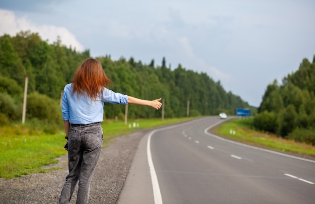 Dziewczyna Zatrzymuje Samochód Na Autostradzie Ręką. Stylowa Kobieta Na Drodze Zatrzymuje Samochód I Rusza W Podróż. Droga W środku Lasu.