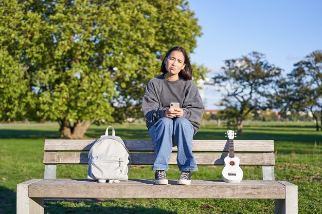 Dziewczyna ze smutną twarzą siedząca na ławce ze smartfonem i ukulele, wyglądająca na zdenerwowaną i rozczarowaną istotą