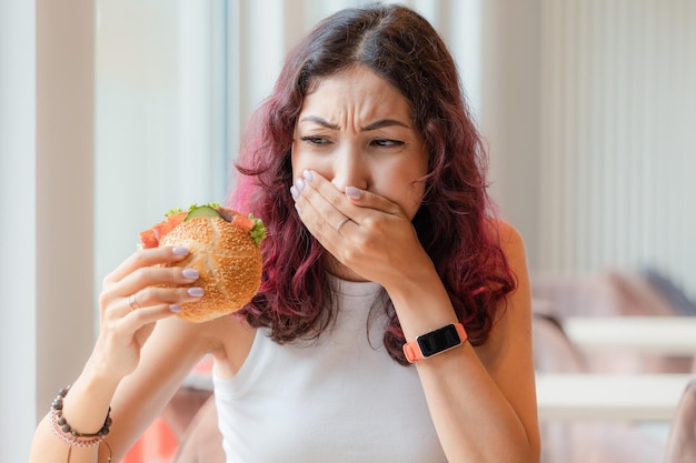 Dziewczyna zjada burgera w kawiarni typu fast food i odczuwa nudności i zgagę Koncepcja niezdrowej żywności i zatrucia pokarmowego