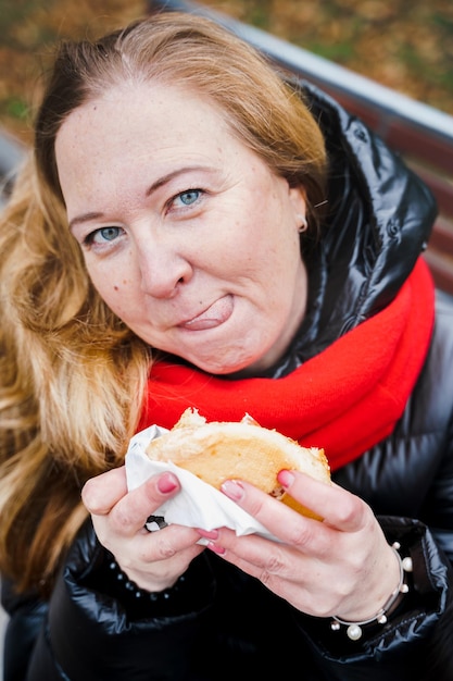 Dziewczynakobieta je dużego hamburgeracheeseburgerŚmieciowe tłuste smaczne uliczne jedzenieBurger na wynosPrzerwa na lunch na świeżym powietrzu w parku siedząc na ławceNiezdrowa dietaWeekendowy relaksPozytywne ciałoProblemy z nadwagą