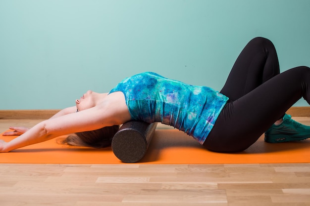 Dziewczynka fitness wykonująca rozluźnianie mięśniowo-powięziowe za pomocą rolki pianki leżącej na podłodze w domu Relaksacja mięśni po wysiłku Zabieg do wykonania w domu