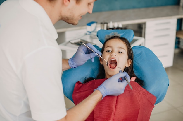 Dziewczynka siedzi w fotelu stomatologicznym z otwartymi ustami, podczas gdy jej dentysta dziecięcy robi badanie zębów.