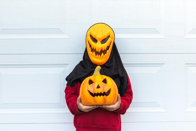 Zdjęcie dziewczynka ubrana w straszną maskę dyniową halloween trzymającą dynię nad białymi drzwiami garażowymi koncepcja uroczystości kostium karnawałowy terror strach i jesień