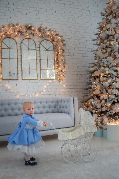 Dziewczynka ubrana w uroczą sukienkę i opaskę, nosi wózek w świątecznie udekorowanym pokoju z girlandą świateł.