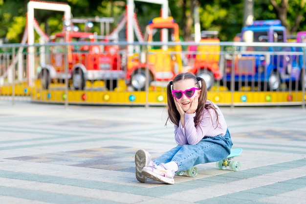 Dziewczynka w parku rozrywki latem jeździ na deskorolce i uśmiecha się ze szczęścia w pobliżu karuzeli w okularach przeciwsłonecznych koncepcja wakacji i wakacji szkolnych